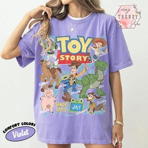 May include: A violet short-sleeved t-shirt featuring a graphic print of Toy Story characters including Woody, Buzz Lightyear, Jessie, Rex, Hamm, Bullseye, Slinky Dog, and an Alien. The text reads Toy Story, Since 1995, and Hi! Lilypad.