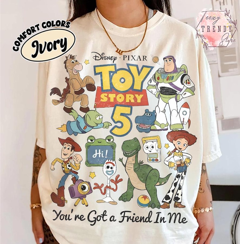 May include: An ivory t-shirt featuring a graphic print of Disney Pixar Toy Story 5 characters including Woody, Buzz Lightyear, Jessie, Rex, Bullseye, and Forky. The text reads Toy Story 5 and You're Got a Friend In Me with a small frog sign saying Hi.