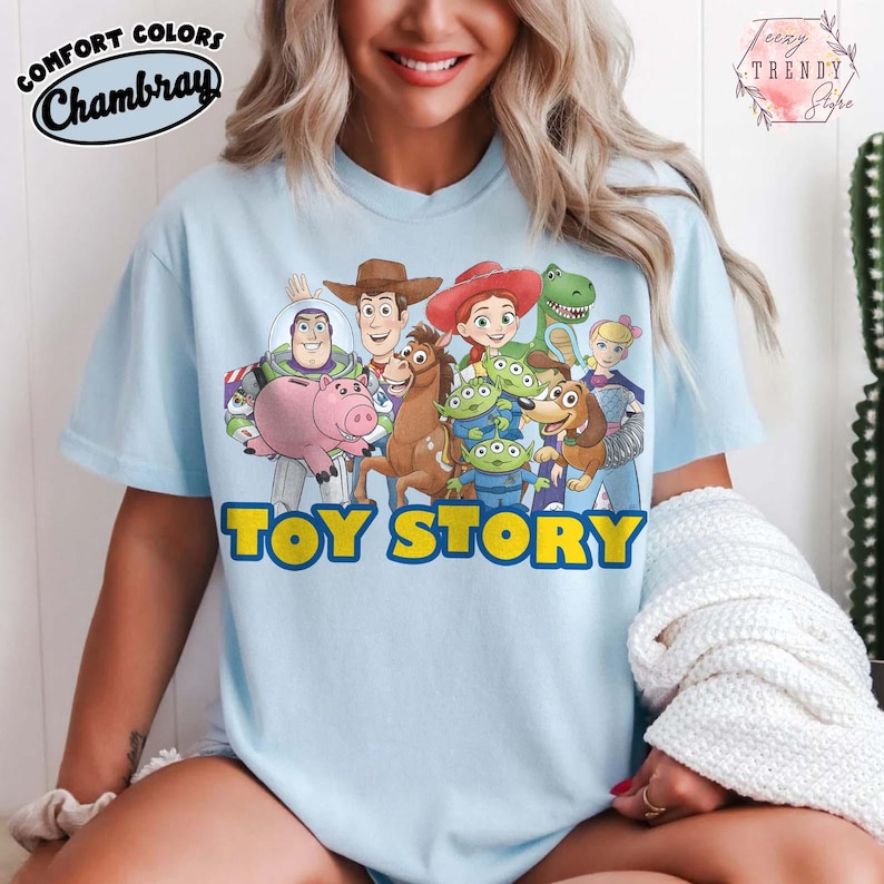 Puede incluir: Camiseta azul claro con un gr&aacute;fico colorido de los personajes de Toy Story, incluyendo a Buzz Lightyear y Woody. Las palabras "TOY STORY" est&aacute;n impresas en amarillo. La camiseta tambi&eacute;n tiene las palabras "Comfort Colors Chambray" impresas en la parte superior izquierda.