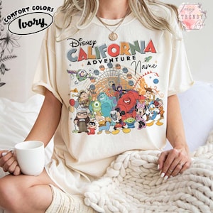 以下が含まれることがあります： 「ディズニー・カリフォルニア・アドベンチャー」の文字と、ディズニーキャラクターのカラフルなグラフィックがプリントされたアイボリー色のTシャツ。半袖でゆったりとしたフィット感。左上に「Comfort Colors」と「Ivory」の文字がプリントされています。