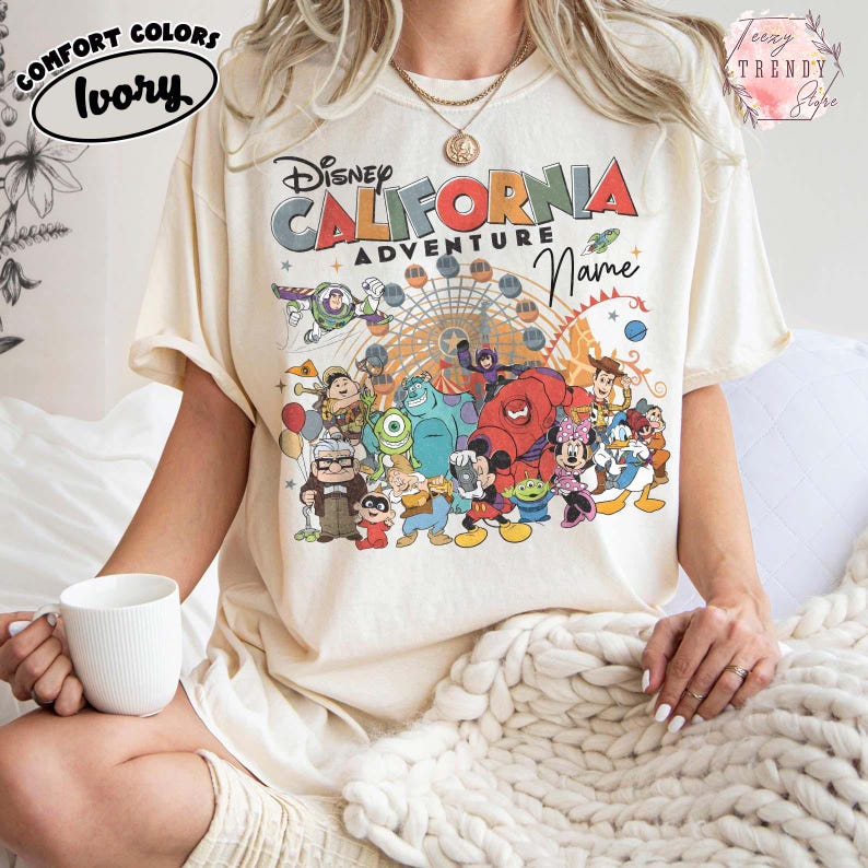 以下が含まれることがあります： 「ディズニー・カリフォルニア・アドベンチャー」の文字と、ディズニーキャラクターのカラフルなグラフィックがプリントされたアイボリー色のTシャツ。半袖でゆったりとしたフィット感。左上に「Comfort Colors」と「Ivory」の文字がプリントされています。