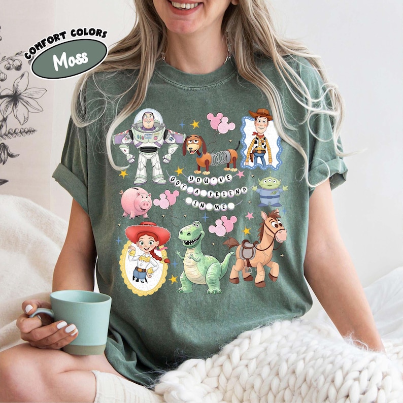 May include: Moss green t-shirt featuring colorful cartoon characters from the Toy Story movie. The shirt includes Buzz Lightyear, Woody, Jessie, and other characters, with the phrase "You've Got a Friend in Me."