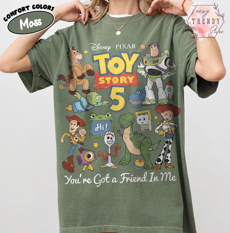 May include: A moss green t-shirt featuring a graphic print of Disney Pixar Toy Story characters including Woody, Buzz Lightyear, Jessie, Rex, Forky, Bullseye, and an alien. The text reads Toy Story 5 and You've Got a Friend In Me.