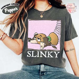 May include: Dark grey Comfort Colors t-shirt with a cartoon image of Slinky Dog from Toy Story. The graphic is in a pink square and the word "SLINKY" is in white block letters.