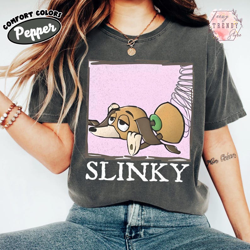 May include: Dark grey Comfort Colors t-shirt with a cartoon image of Slinky Dog from Toy Story. The graphic is in a pink square and the word "SLINKY" is in white block letters.
