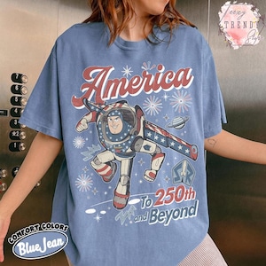 Puede incluir: Una camiseta de color azul vaquero con un gr&aacute;fico de un personaje de guardi&aacute;n espacial con un traje rojo, blanco y azul. El texto dice America, To 250th and Beyond, e incluye ilustraciones de fuegos artificiales, un planeta y un cohete.