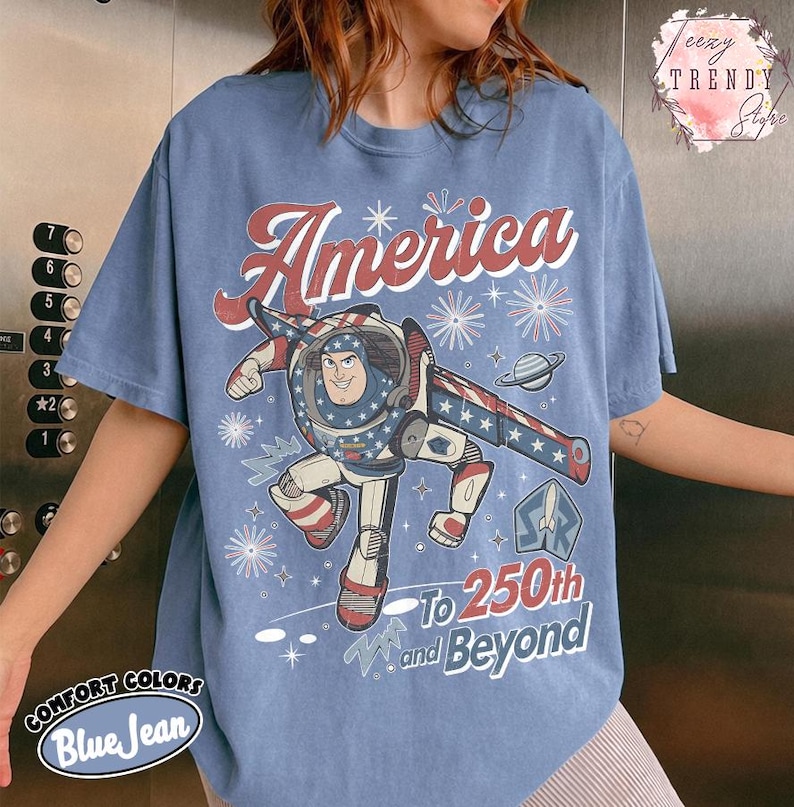 Puede incluir: Una camiseta de color azul vaquero con un gr&aacute;fico de un personaje de guardi&aacute;n espacial con un traje rojo, blanco y azul. El texto dice America, To 250th and Beyond, e incluye ilustraciones de fuegos artificiales, un planeta y un cohete.