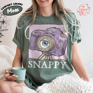 May include: A moss green t-shirt featuring a purple camera design with an eye-shaped lens and the word "SNAPPY" below. The shirt also has the words "COMFORT COLORS" and "Moss" printed on it.