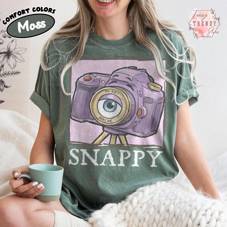 May include: A moss green t-shirt featuring a purple camera design with an eye-shaped lens and the word "SNAPPY" below. The shirt also has the words "COMFORT COLORS" and "Moss" printed on it.
