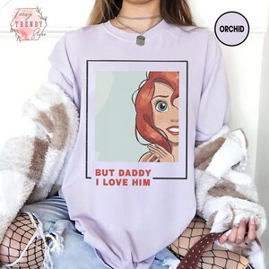 Puede incluir: Una sudadera lila con una ilustraci&oacute;n enmarcada de un personaje de dibujos animados con pelo rojo y ojos azules. El texto "BUT DADDY I LOVE HIM" est&aacute; impreso debajo. La sudadera tiene mangas largas.