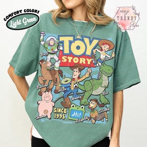 May include: A light green t-shirt featuring a large graphic print of Toy Story characters including Buzz Lightyear, Woody, Jessie, Bullseye, Rex, Hamm, Slinky, and an Alien. The text reads Toy Story, Since 1995, and Hi! on a green frog-shaped sign.