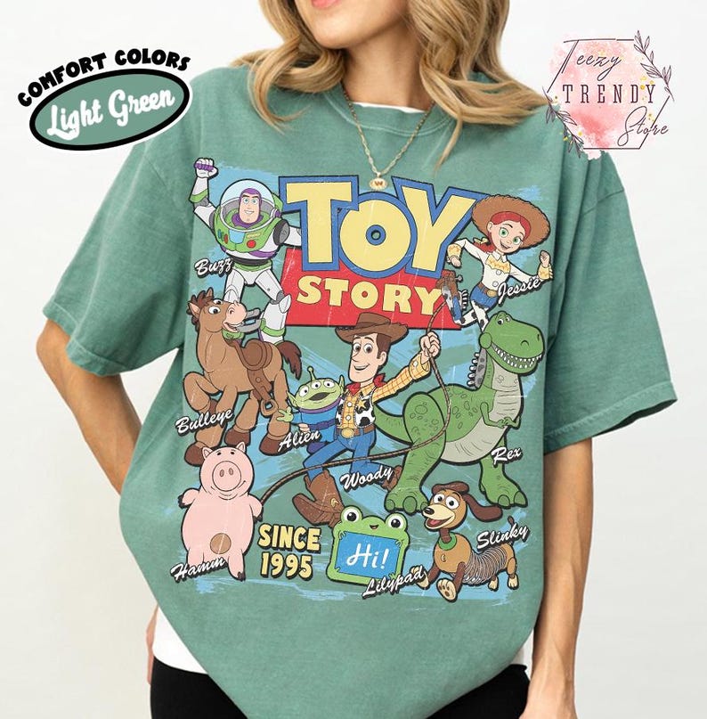 May include: A light green t-shirt featuring a large graphic print of Toy Story characters including Buzz Lightyear, Woody, Jessie, Bullseye, Rex, Hamm, Slinky, and an Alien. The text reads Toy Story, Since 1995, and Hi! on a green frog-shaped sign.
