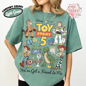 May include: A light green short-sleeve t-shirt featuring a graphic print of Toy Story characters including Woody, Buzz Lightyear, Jessie, Rex, and Forky. The text reads Toy Story 5 and You've Got a Friend In Me. The shirt has a vintage-style design.
