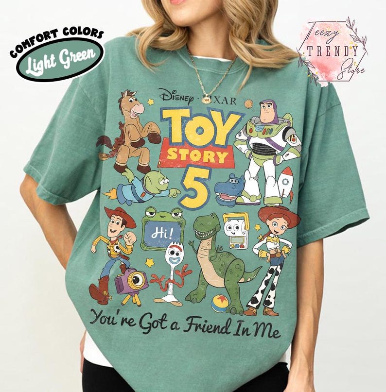 May include: A light green short-sleeve t-shirt featuring a graphic print of Toy Story characters including Woody, Buzz Lightyear, Jessie, Rex, and Forky. The text reads Toy Story 5 and You've Got a Friend In Me. The shirt has a vintage-style design.