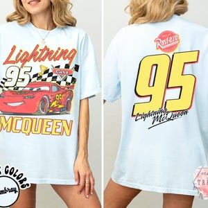 May include: Light blue t-shirt featuring Lightning McQueen from the Cars movie. The front graphic shows McQueen with the number 95, a checkered flag, and the text "Lightning McQueen". The back displays a large number 95 and the text "Lightning McQueen".
