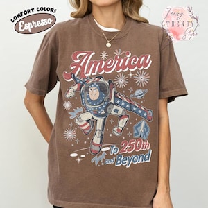 Puede incluir: Una camiseta de manga corta color marr&oacute;n espresso con un gr&aacute;fico de un personaje de ranger espacial en un traje rojo, blanco y azul. El texto dice America To 250th and Beyond, junto a ilustraciones de fuegos artificiales y estrellas.