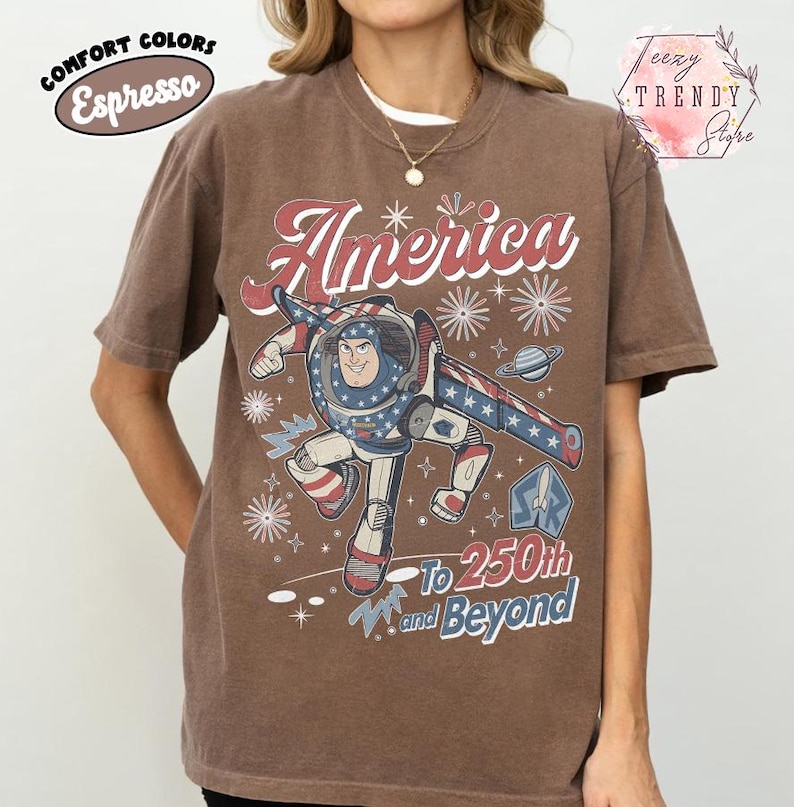 Puede incluir: Una camiseta de manga corta color marr&oacute;n espresso con un gr&aacute;fico de un personaje de ranger espacial en un traje rojo, blanco y azul. El texto dice America To 250th and Beyond, junto a ilustraciones de fuegos artificiales y estrellas.