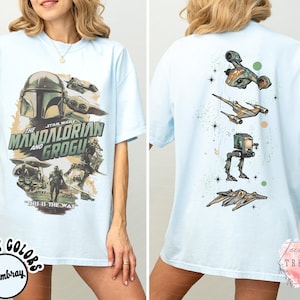 K&ouml;nnte beinhalten: Ein hellblaues T-Shirt mit einem Star Wars-Motiv auf der Vorderseite, das The Mandalorian und Grogu zeigt, inklusive des Textes The Mandalorian and Grogu und This is the Way. Die R&uuml;ckseite zeigt eine vertikale Anordnung von vier Raumfahrzeugen.