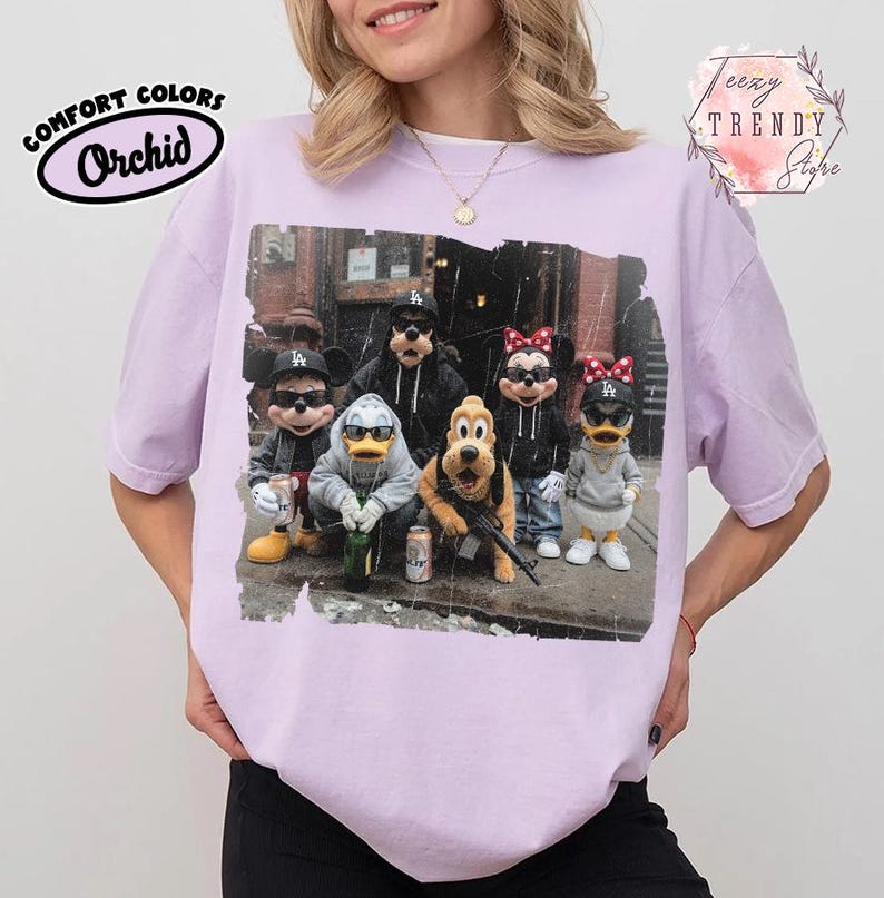 May include: A light purple t-shirt featuring a graphic print of cartoon characters wearing black baseball caps and sunglasses. The design shows Mickey, Donald, Goofy, Pluto, Minnie, and Daisy in a street setting with cans and a rifle.