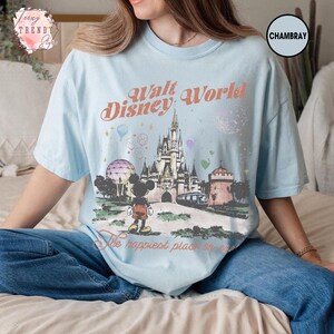 May include: Light blue t-shirt featuring a graphic of the Walt Disney World castle, Mickey Mouse, and the text "Walt Disney World" and "The happiest place on earth." The shirt is made of a soft material.