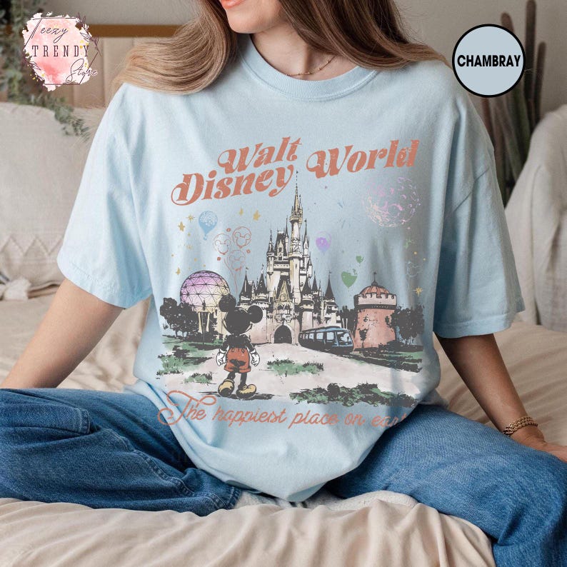 May include: Light blue t-shirt featuring a graphic of the Walt Disney World castle, Mickey Mouse, and the text "Walt Disney World" and "The happiest place on earth." The shirt is made of a soft material.