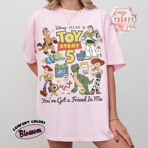 May include: A light pink t-shirt featuring a colorful graphic print of Disney Pixar Toy Story characters including Woody, Buzz Lightyear, Jessie, Rex, and Forky. The text reads Toy Story 5 and You've Got a Friend In Me. The shirt has short sleeves and a crew neck.