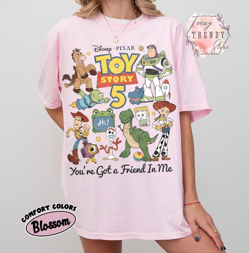 May include: A light pink t-shirt featuring a colorful graphic print of Disney Pixar Toy Story characters including Woody, Buzz Lightyear, Jessie, Rex, and Forky. The text reads Toy Story 5 and You've Got a Friend In Me. The shirt has short sleeves and a crew neck.