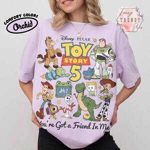 May include: A light purple short-sleeve t-shirt featuring a graphic print of Disney Pixar Toy Story characters including Woody, Buzz Lightyear, Jessie, Rex, and Forky. The text reads Toy Story 5 and You've Got a Friend In Me with a small Hi sign.