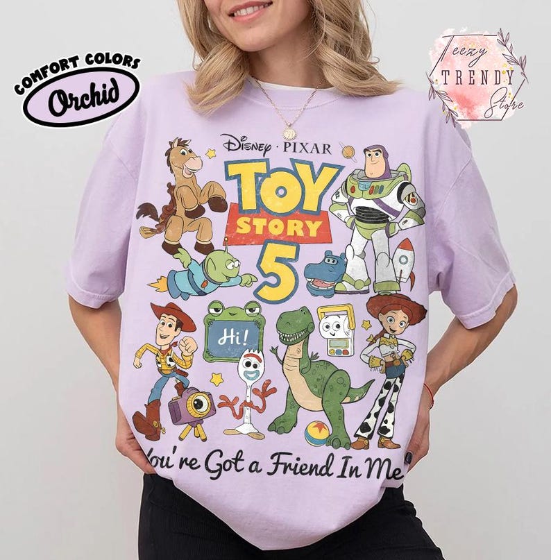 May include: A light purple short-sleeve t-shirt featuring a graphic print of Disney Pixar Toy Story characters including Woody, Buzz Lightyear, Jessie, Rex, and Forky. The text reads Toy Story 5 and You've Got a Friend In Me with a small Hi sign.
