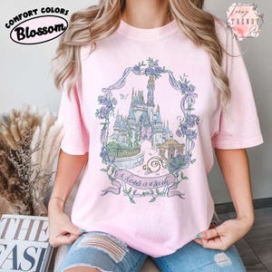 Puede incluir: Camiseta rosa claro con un gr&aacute;fico floral de un castillo. El gr&aacute;fico presenta un castillo, un carruaje y el texto "a dream is a wish". La camiseta tiene los logotipos Comfort Colors y Blossom.