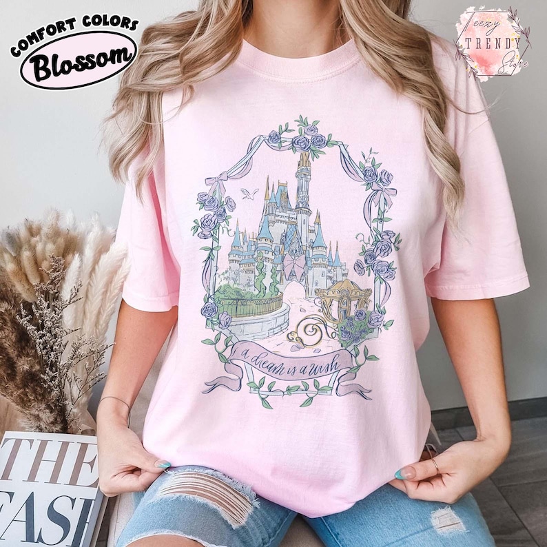 Puede incluir: Camiseta rosa claro con un gr&aacute;fico floral de un castillo. El gr&aacute;fico presenta un castillo, un carruaje y el texto "a dream is a wish". La camiseta tiene los logotipos Comfort Colors y Blossom.