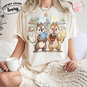 May include: Ivory-colored t-shirt featuring a cartoon illustration of Chip and Dale holding treats in front of a theme park backdrop. The shirt has the words "Comfort Colors" and "Ivory" printed on it. The shirt is a relaxed fit.