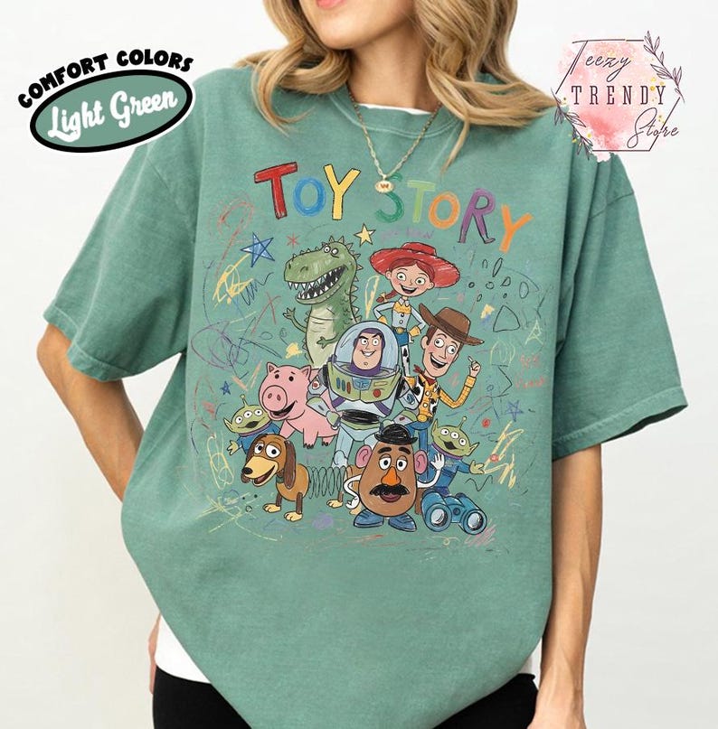 May include: Light green t-shirt featuring the words "Toy Story" in colorful letters, with cartoon characters from the movie. The shirt also has the words "Comfort Colors" and "Light Green" in a circular design.