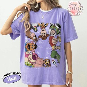 May include: A violet Comfort Colors t-shirt featuring a colorful graphic of Toy Story characters. The design includes Woody, Buzz Lightyear, and other characters from the animated film. The shirt also has the text "Comfort Colors Violet".