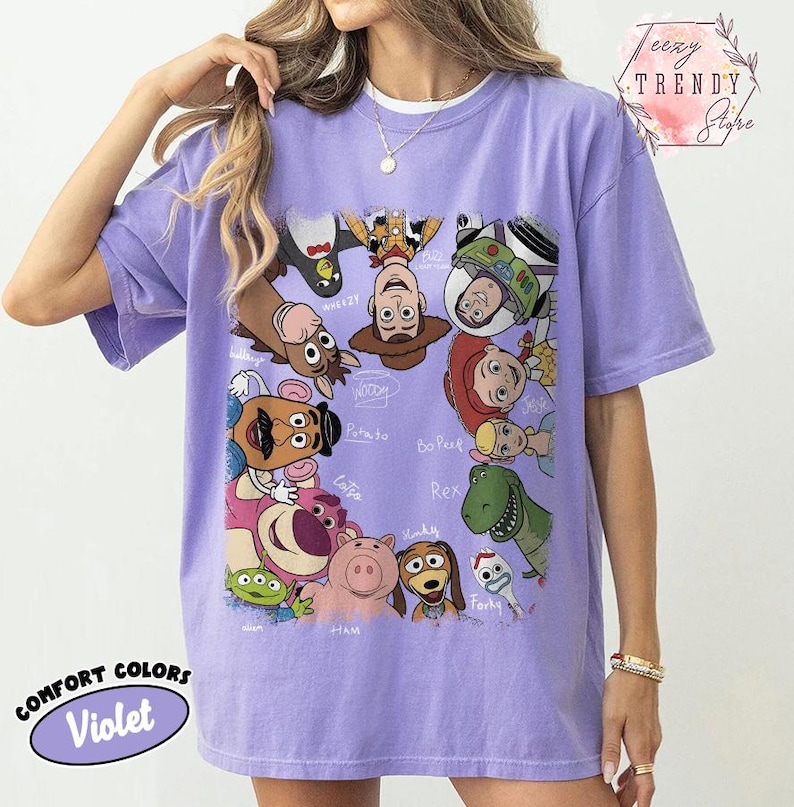 May include: A violet Comfort Colors t-shirt featuring a colorful graphic of Toy Story characters. The design includes Woody, Buzz Lightyear, and other characters from the animated film. The shirt also has the text "Comfort Colors Violet".