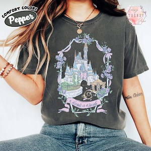 Puede incluir: Camiseta gris oscuro con un gr&aacute;fico colorido de un castillo, rodeado de flores y cintas. El texto "a dream is a wish" est&aacute; debajo del castillo. La camiseta tiene los logotipos Comfort Colors y Teezy Trendy Style.