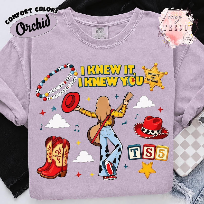 May include: A light purple t-shirt featuring a graphic of a person with long hair playing a guitar, surrounded by cowboy boots, hats, a beaded bracelet with the text 'you've got a friend in me', a sheriff badge, and the text 'I KNEW IT, I KNEW YOU'.