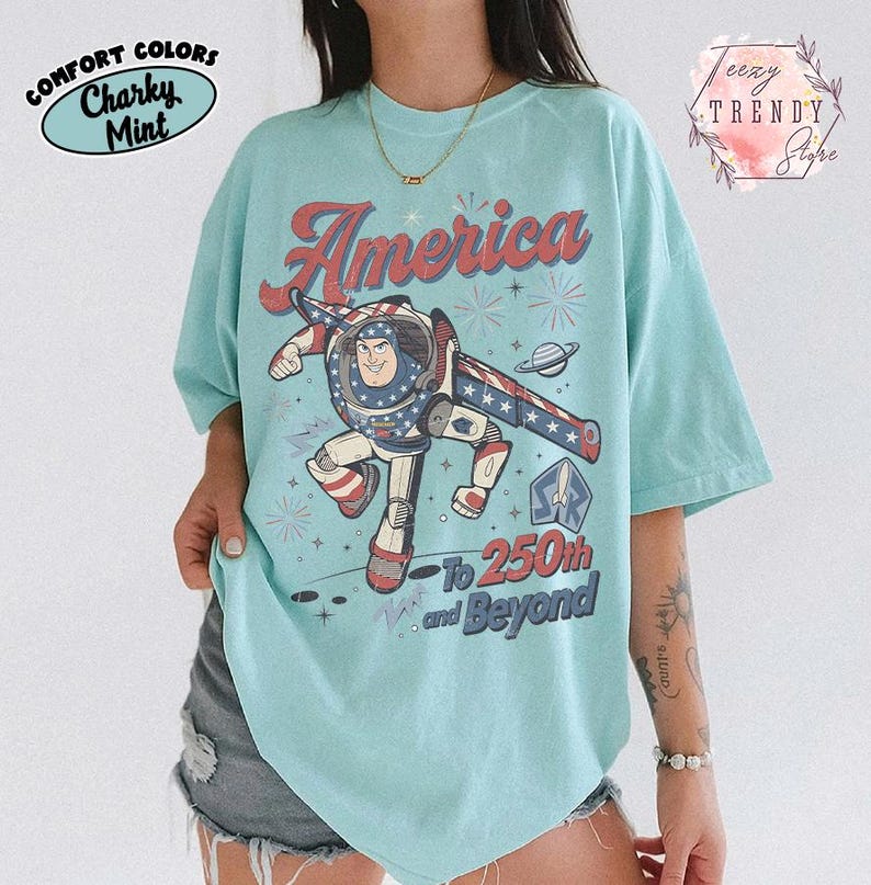Puede incluir: Una camiseta de manga corta color verde menta con un gr&aacute;fico de un personaje de guardi&aacute;n espacial en pose de vuelo. El texto dice America To 250th and Beyond, acompa&ntilde;ado de ilustraciones de un planeta, estrellas y fuegos artificiales en rojo, blanco y azul.
