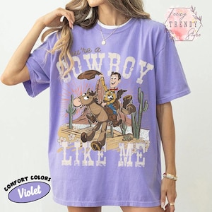May include: A violet short-sleeved t-shirt featuring a graphic print of a cowboy riding a horse in a desert landscape with cacti. The text "You're a COWBOY LIKE ME" is printed in a distressed white font above and below the central illustration.
