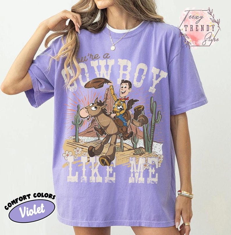 May include: A violet short-sleeved t-shirt featuring a graphic print of a cowboy riding a horse in a desert landscape with cacti. The text "You're a COWBOY LIKE ME" is printed in a distressed white font above and below the central illustration.