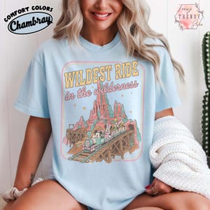 Może przedstawiać: Jasnoniebieska koszulka z grafiką retro przedstawiającą pociąg na drewnianym moście na tle pasma g&oacute;rskiego. Tekst "WILDEST RIDE in the wilderness" jest wyświetlany w stylu vintage. Koszulka ma r&oacute;wnież napis "Comfort Colors Chambray".