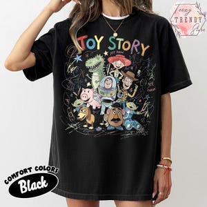 May include: Black Comfort Colors t-shirt featuring the words "TOY STORY" above a colorful graphic of characters from the animated film. The design includes Woody, Buzz Lightyear, and other characters. The shirt has a crew neck.
