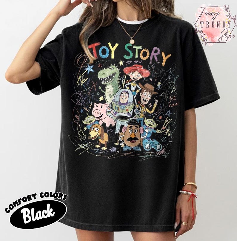 May include: Black Comfort Colors t-shirt featuring the words "TOY STORY" above a colorful graphic of characters from the animated film. The design includes Woody, Buzz Lightyear, and other characters. The shirt has a crew neck.