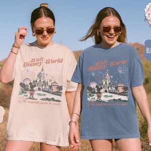 May include: Two women wearing ivory and blue jean-colored t-shirts. Each shirt features a graphic of Cinderella's castle, Mickey Mouse, and the text "Walt Disney World" and "The happiest place on earth."