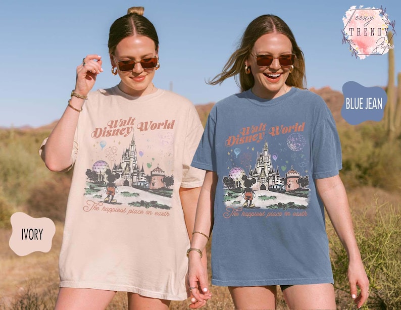 May include: Two women wearing ivory and blue jean-colored t-shirts. Each shirt features a graphic of Cinderella's castle, Mickey Mouse, and the text "Walt Disney World" and "The happiest place on earth."