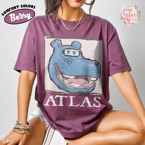 May include: Berry-coloured t-shirt featuring a blue hippo graphic and the word "ATLAS" in white. The shirt has short sleeves and a crew neck. The words "Comfort Colors" and "Berry" are printed on the upper left.