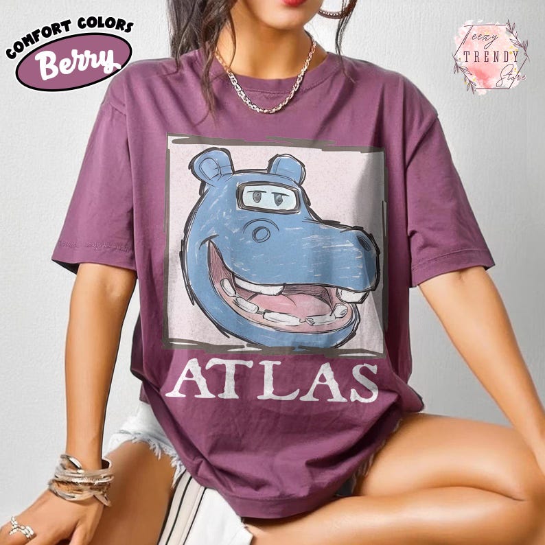 May include: Berry-coloured t-shirt featuring a blue hippo graphic and the word "ATLAS" in white. The shirt has short sleeves and a crew neck. The words "Comfort Colors" and "Berry" are printed on the upper left.