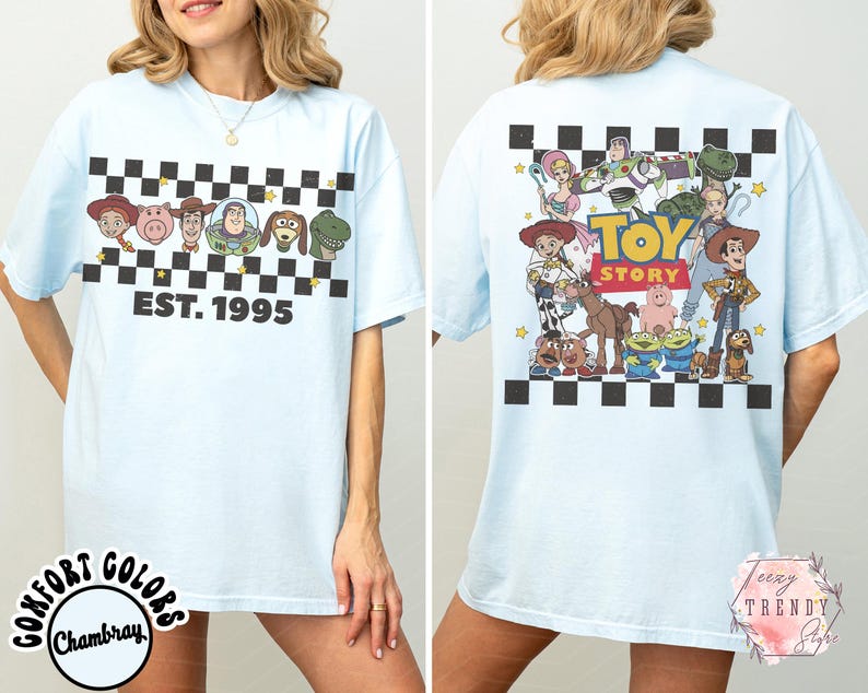 Op de afbeelding: Lichtblauw T-shirt met personages uit de film Toy Story. De voorkant heeft een zwart-wit geblokt patroon met de tekst "EST. 1995" en karakterillustraties. De achterkant heeft een grotere Toy Story-afbeelding.