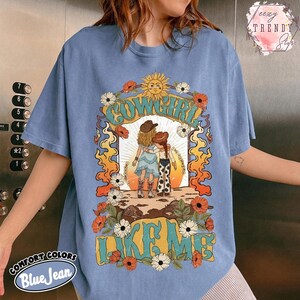 May include: A blue jean colored t-shirt featuring a graphic print of two figures in cowboy hats standing together. The design includes a sun, flowers, and the text "COWGIRL LIKE ME" in a stylized font. The shirt has short sleeves and a crew neckline.
