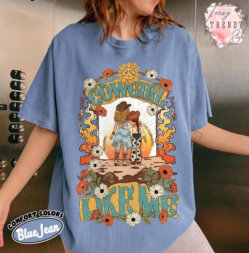 May include: A blue jean colored t-shirt featuring a graphic print of two figures in cowboy hats standing together. The design includes a sun, flowers, and the text "COWGIRL LIKE ME" in a stylized font. The shirt has short sleeves and a crew neckline.